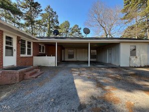 A house with a brick and siding exterior features a large carport and several windows. Two satellite dishes are mounted on the roof. The surrounding area includes trees and a driveway with gravel and concrete.