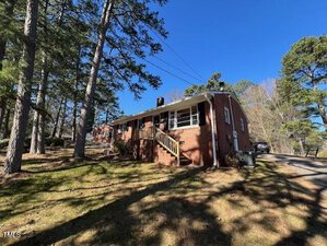 A single-story, red brick house with a small front porch and a few steps leading up to the entrance. Surrounded by tall trees, the property has a sloped lawn and a paved driveway on the right. The sky is clear and sunny.