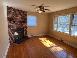 A living room with a brick fireplace, wooden floor, and ceiling fan. A large window lets in natural light, and there's a smaller window with a blind partially open. The walls are painted in a cream color.