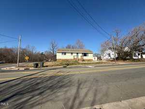 A single-story beige house with a gray roof is situated on a corner lot. The house is bordered by a road with double yellow lines. Leafless trees surround the property, and utility lines cross the clear blue sky.