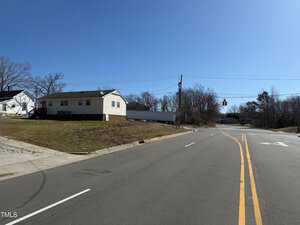 A residential area with a single-story house on a slightly elevated grass plot is on the left. The road curves to the right with neat lane markings. Traffic lights and leafless trees are visible in the background under a clear blue sky.