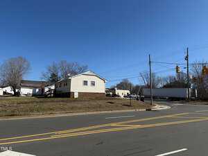 A small, single-story house with a light-colored exterior sits on a grassy lot at the corner of an intersection. A large truck is also visible on the road. Leafless trees are in the background under a clear blue sky.