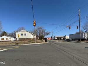 An intersection with traffic lights on a clear day. A two-story house is on the left, and an industrial building with a truck is on the right. Power lines and a road sign are visible. The sky is clear and blue.