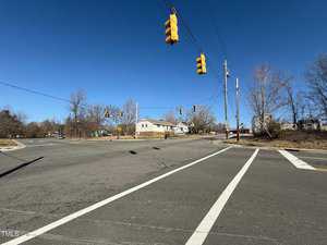 A clear, sunny day at a mostly empty intersection with traffic lights hanging overhead. The road is marked with crosswalk lines. Bare trees and a few houses are visible in the background under a bright blue sky.