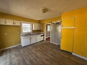 A small, worn kitchen with yellow walls and white cabinets. There is a stainless steel sink with a window above it, a dishwasher to the right, and wood flooring. A doorway leads to another room, and there is a ceiling light fixture.