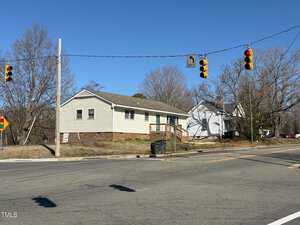 Intersection with traffic signals showing a red light. A residential area in the background features a single-story house with light siding and a porch. Leafless trees and a clear sky are visible. There's a trash bin at the corner.