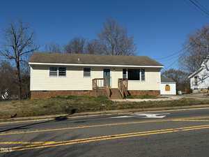 A single-story, beige house with a gray roof and white trim sits behind a two-lane road. The house has a front porch with stairs and railing. Sparse grass and leafless trees are visible in the yard and surroundings under a clear blue sky.
