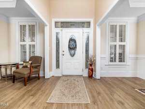 A spacious entryway features a white front door with an oval glass insert, flanked by sidelights. There are wooden floors, a patterned rug, a decorative vase, and an armchair with a small side table. Shuttered windows are on either side.