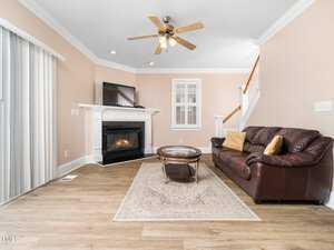 Living room with a brown leather sofa, glass coffee table, and rug on a wooden floor. A fireplace with a TV on the mantel is in the corner. Ceiling fan with lights overhead. Window with blinds and stairs leading upward visible in the background.