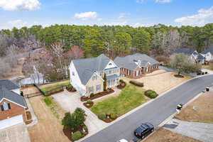 Aerial view of a suburban neighborhood showing two large houses with well-maintained lawns, driveways, and landscaping. The street curves alongside, and there are trees and greenery in the background under a partly cloudy sky.
