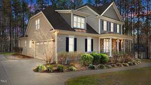 A two-story house at 1196 Smith Creek Way, with beige siding, white trim, and dark shutters is surrounded by landscaped bushes and a lawn. Located in Wake Forest, it boasts a gabled roof and a porch. The backdrop features trees against a sky transitioning from blue to pink.
