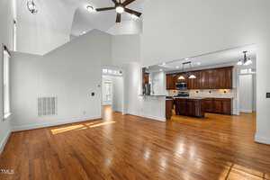 A spacious, well-lit room with wooden flooring connects to an open kitchen featuring dark wood cabinets, stainless steel appliances, and a light granite countertop. Recessed and pendant lighting adorn the ceiling. Natural light filters in from large windows.