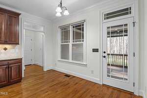 Kitchen area with hardwood floors, white walls, and a ceiling light fixture. A wooden cabinet is partially visible on the left. There is a window and glass door leading to a deck outside, with trees in the background.
