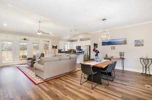 Open-concept living and dining area with large windows. Features a gray sectional sofa, armchair, area rug, and ceiling fan. Dining area has a wooden table with black chairs and a modern light fixture. Brown hardwood floors throughout.