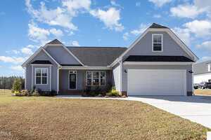 A light gray single-story house with white trim and a black roof is seen under a blue sky with clouds. The house features a two-car garage, a front porch with columns, and a neatly trimmed lawn.