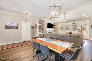 A modern living and dining area with a wooden table, six chairs, and a sofa facing a TV on a mantel. Shelves display decor items. A large window and glass doors provide natural light. A geometric light fixture hangs above the table. Wood flooring throughout.