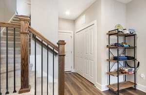 A hallway featuring a staircase with wooden handrails and carpeted steps on the left. There is a white door at the end of the hallway and a closet with double doors. A wooden shelf holds books and electronic equipment on the right.