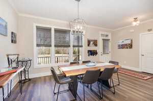 A dining room with a wooden table and six black chairs. A geometric chandelier hangs above the table. Large windows with blinds let in natural light. Walls are beige, and the floor is wooden. Various framed photos are on the walls.