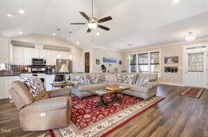 A spacious living room with an open kitchen layout. The room has a large beige sectional sofa, a brown recliner, a patterned red rug, and a ceiling fan. The kitchen area has white cabinets and stainless steel appliances. Wall art and decor are visible.