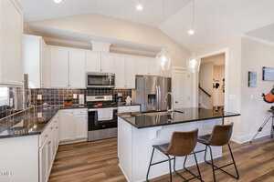 Modern kitchen with white cabinetry, black granite countertops, and stainless steel appliances. Two brown stools sit at the island, beneath two pendant lights. The backsplash features dark tiles, and the floor has wood-style planks.