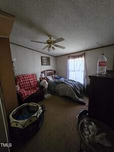 A dimly lit bedroom features a bed with rumpled covers, a ceiling fan, a red armchair with a patterned throw, a pet playpen, and a dresser with a large bottle on top. A curtain-draped window lets in some light. The floor is carpeted.