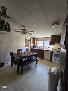 A kitchen with a wooden dining table and four chairs. The room has a patterned wallpaper, a ceiling fan, and large windows. The kitchen features dark wood cabinets, a white stove, and a sink with dishes. There are items on shelves and the floor is tiled.