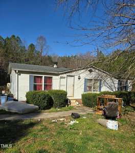 A small, single-story house with light-colored siding and a dark roof. There are red curtains on one window and a bush-lined pathway leading to the entrance. The yard has some scattered items and is bordered by trees in the background.