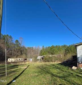 A grassy yard with scattered debris and old trailers is bordered by a line of trees. A tilted utility pole and overhead wires are visible. The sky is clear and blue.