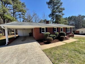 Nestled on 1723 Lynne Avenue in Henderson, this brick single-story house boasts a gray roof and carport. It features a chimney, blue shutters, and a charming front garden with shrubs. A concrete driveway guides you to the carport, all framed by tall trees in the background.