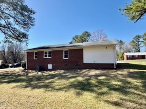 Single-story house with a red brick exterior and white siding on one side. The house has several small windows and is surrounded by a grassy lawn. Trees are visible in the background under a clear blue sky.