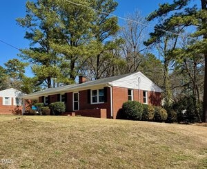 A one-story brick house with white trim is surrounded by trees and bushes. The front yard has a slight slope and is covered in grass. A clear blue sky is visible overhead.