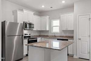 Modern kitchen with white cabinets, stainless steel appliances including a refrigerator, oven, and microwave. Features a granite-topped island with a sink. Overhead lighting and a small window provide additional illumination. White door to the right.