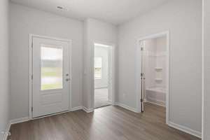 A minimalist hallway with light wood flooring features a frosted glass door on the left, an open door to a carpeted room in the center, and an open door to a bathroom with a white bathtub and shelves on the right. Walls are painted white.