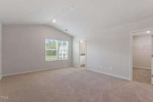 An empty room with beige carpet and light gray walls. A large window with blinds is on the left wall. Doorways to a bathroom and a walk-in closet are on the right. The ceiling is vaulted with recessed lighting.