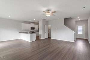Spacious, unfurnished room with light gray walls and wood flooring. A ceiling fan is centered above. The open kitchen features white cabinets, stainless steel appliances, and a marble countertop. A door and staircase are visible in the background.