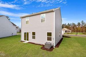 A two-story house with beige siding, large windows, and a glass sliding door on a grassy lawn. An air conditioning unit is positioned near the back wall. A clear blue sky and a neighboring house are visible in the background.