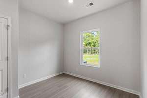 Small, empty room with light gray walls and wood-style flooring. There's a single window revealing a view of green trees outside. A white door is partially visible on the left. Ceiling light is on, brightening the space.