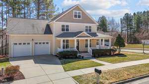 A two-story house with a beige exterior, white trim, and a gabled roof. It has a covered front porch, two-car garage, and a driveway. The lawn is neatly maintained, and there are trees and shrubs around the property. A mailbox is in the foreground.