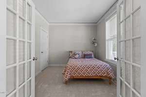 A small bedroom features a wooden bed with a patterned quilt, flanked by two windows with blinds. The room has beige walls and a carpeted floor. Glass-panel double doors open into the space. A closed white door is visible on the left wall.