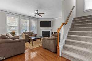 Living room with brown sofas, a wooden coffee table, and a carpet on hardwood flooring. There is a fireplace with a flat-screen TV above it, large windows with blinds, a ceiling fan, and a staircase with carpeted steps on the right.