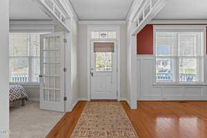 A foyer with a wooden floor and a patterned rug leads to a white front door with a frosted glass window. To the left, a room with a French door and a visible bed. Large windows on each side let in ample light. Walls are painted with white and a deep red accent.