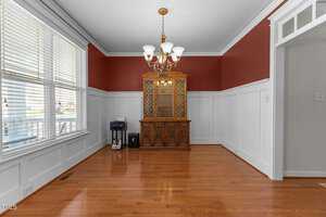 An empty room with white wainscoting and red upper walls. There is a wooden china cabinet, a small printer, and a shredder on the left. The floor is wooden, and a chandelier hangs from the ceiling. Sunlight streams through the large window on the left.