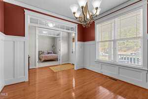 A vacant dining room with polished wooden floors, white wainscoting, and red upper walls. A brass chandelier hangs from the ceiling. Double glass doors open to a room with a bed. Large windows feature blinds, offering a view outside.