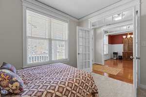 A bedroom with a bed featuring a patterned coverlet and decorative pillows is shown. The room has light-colored walls, a large window with blinds, and an open doorway leading to another room with wood flooring and white paneled walls.