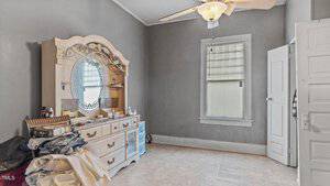 A sparsely furnished room with gray walls features a vintage dresser with an ornate mirror. Piles of clothes and various items are on top. A ceiling fan is above, and a window with blinds lets in light. An open door is on the right.