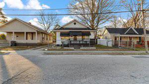 A street view of three small, single-story houses on 123 S College Street. The middle house features a black awning and fenced yard. Light-colored houses with porches flank it, set against bare trees under a clear sky in Henderson. Power lines crisscross above.