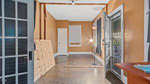 An empty room with tan walls and a white ceiling features a floor lamp, an open doorway leading outside, a covered window, and a closed door. There is a wooden board propped against the wall and a long plank on the floor.
