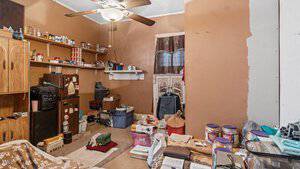A cluttered room with brown walls and a ceiling fan. Various items are scattered, including bins, bags, and storage containers. Shelves with jars and cans line one wall. A small brown fridge and a wooden dresser are also present.