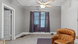 A small room with gray walls and a white ceiling fan. There is an armchair in the foreground, and a window with sheer black curtains in the background. A partially open door is on the left, and there are wires on the floor.