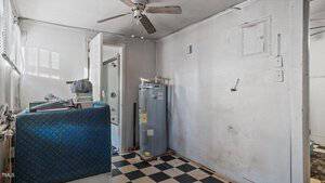 A small, plain room with a black-and-white checkered floor. There's a water heater against the wall, stacks of materials in the corner, and a ceiling fan. A door is partially open, revealing a glimpse of another room.
